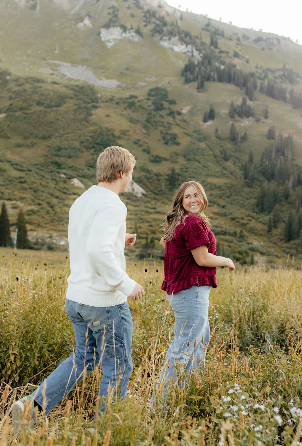 A woman in a red top smiles back at her partner while walking in a mountain field.