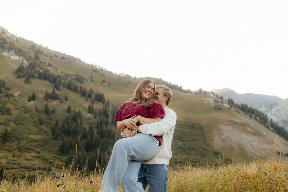 A couple embraces and laughs together while standing in a golden mountain meadow.