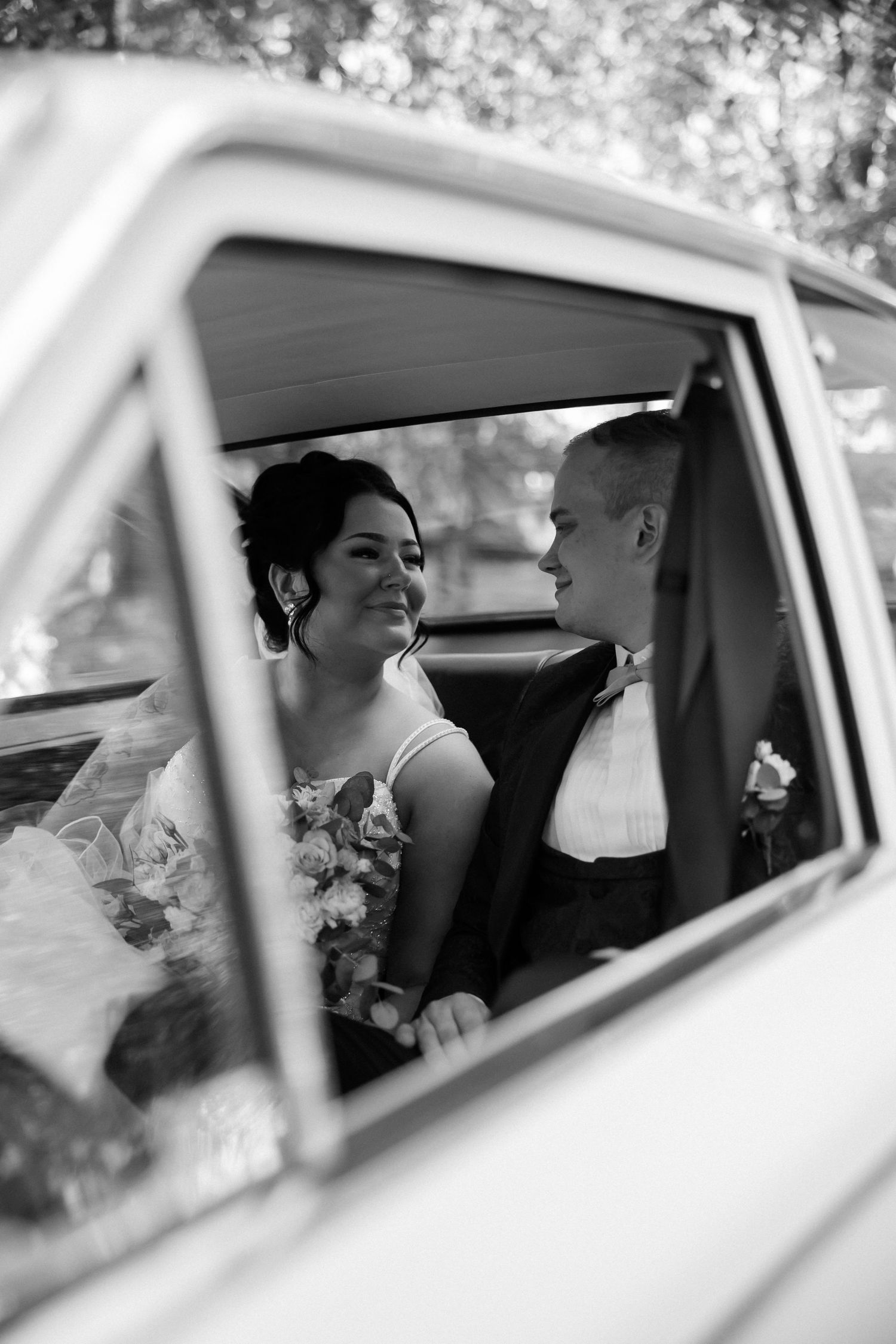 Black and white photo of newlyweds sharing intimate moment through vintage car window.