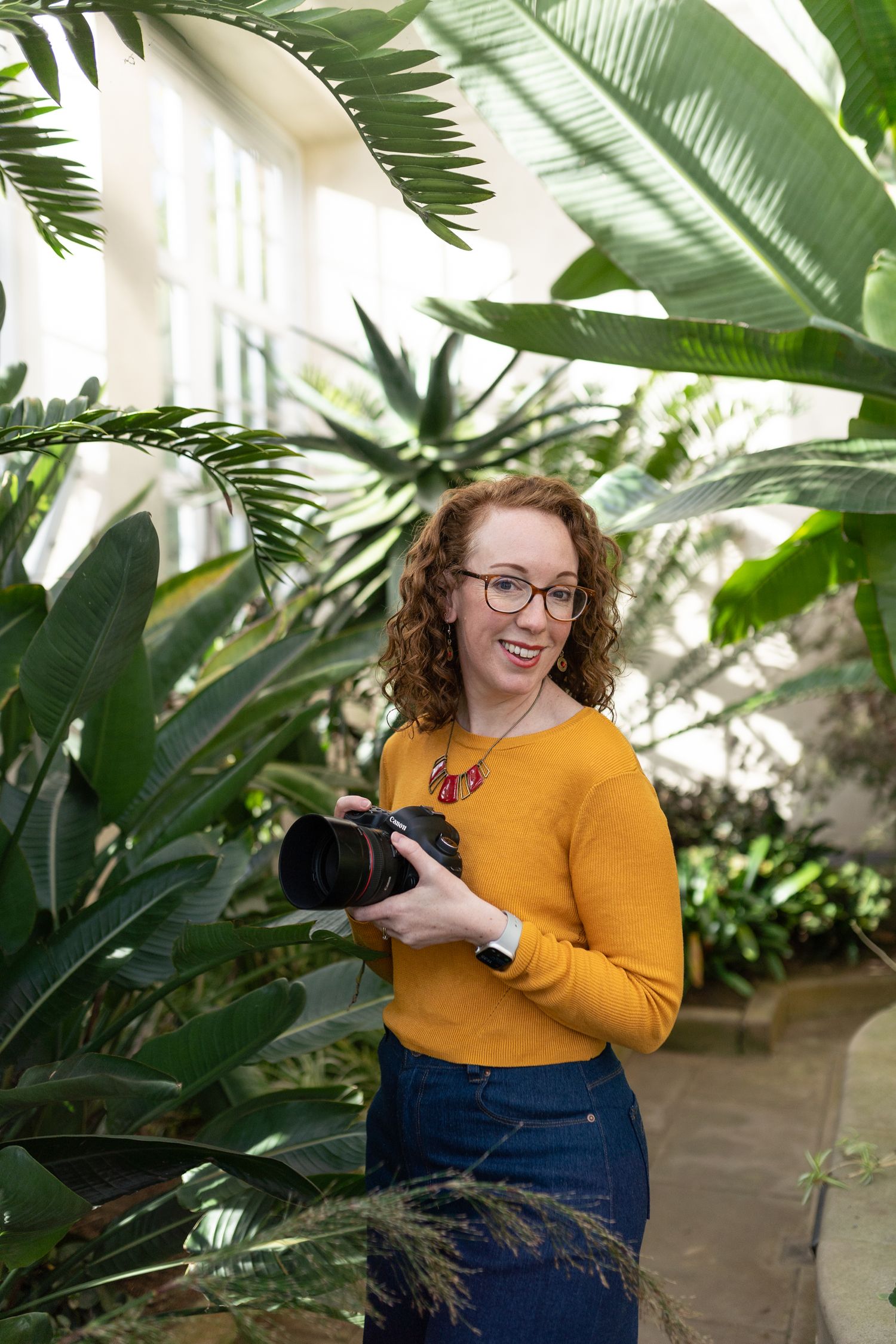 Photographer with a camera stands among tropical plants in a greenhouse setting.