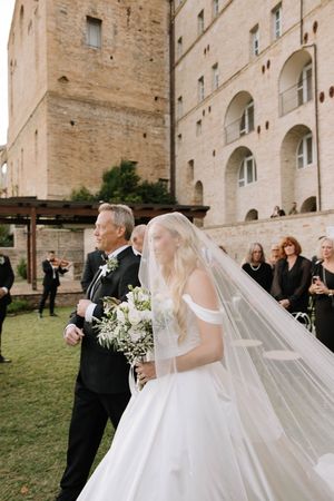 A bride in a flowing white gown with long veil walks outdoors near a stone building.