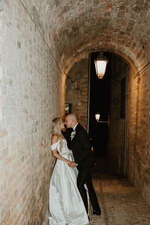 A couple shares an intimate moment in a dimly lit brick tunnel with wall sconces.