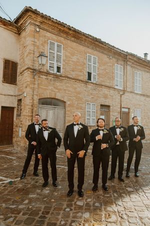 Groomsmen in black tuxedos pose together against a stone building exterior.