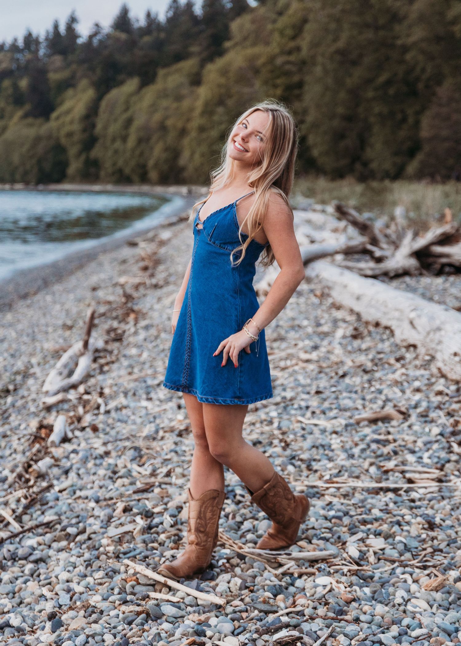 A person wearing a blue dress and brown boots walks along a rocky beach with driftwood and forest in the background.