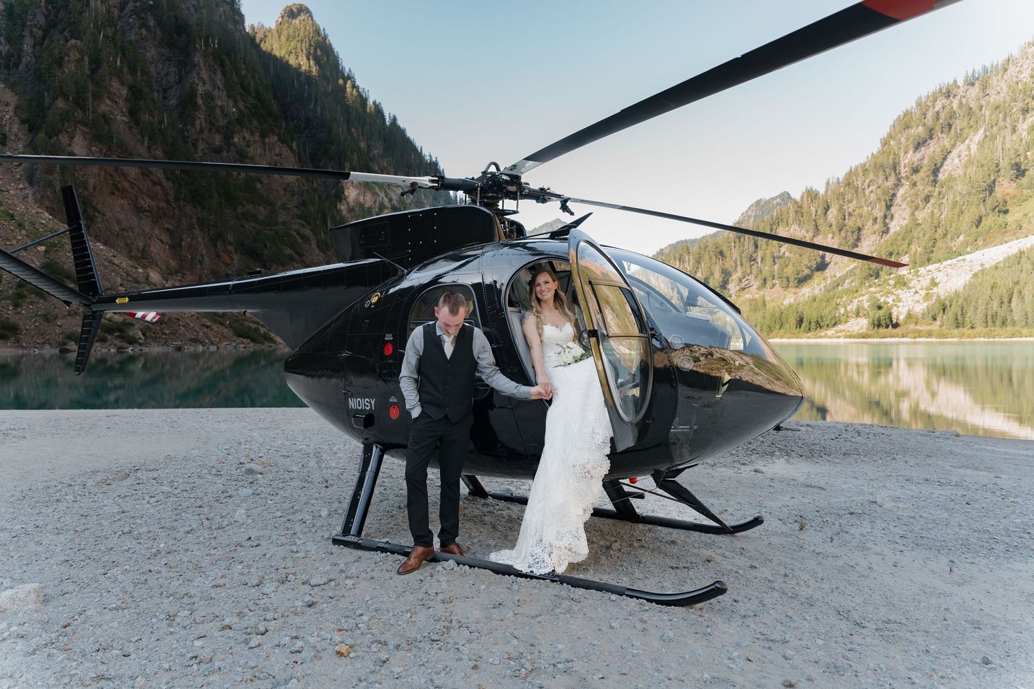 A newlywed couple poses by a black helicopter against a mountainous landscape.