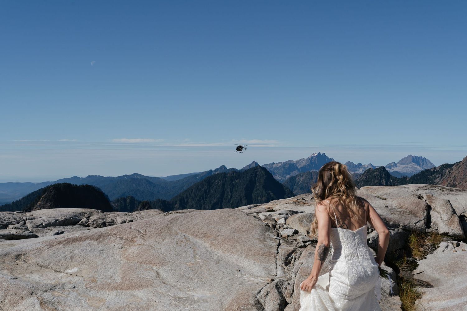 A mountain wedding photoshoot with a helicopter flying in the background against blue skies.