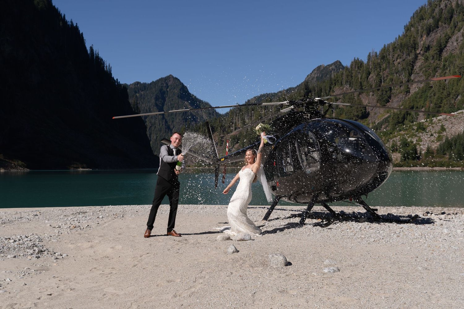 A wedding couple strikes playful poses next to a black helicopter on a mountain shore.