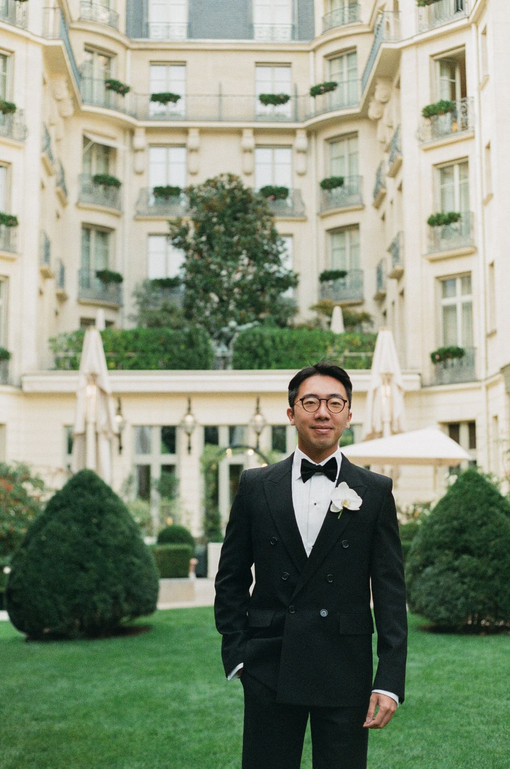 A groomsman in a black tuxedo and bowtie stands on manicured lawn outside an elegant French-style building.