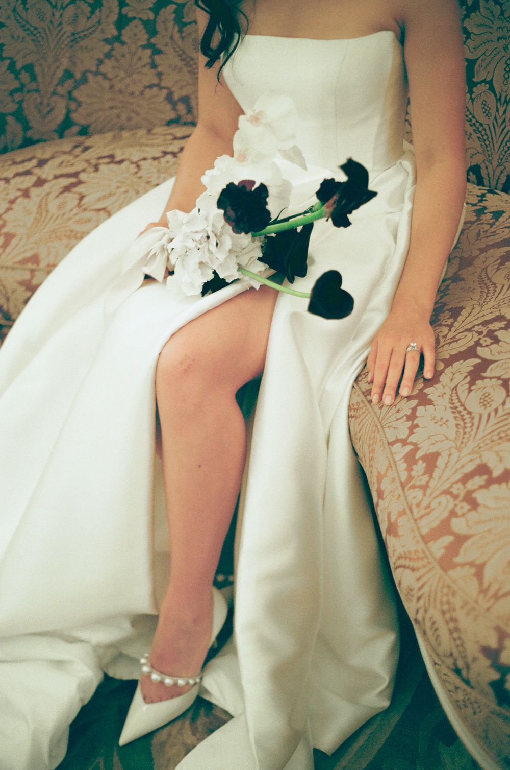 A close-up of a white wedding dress with black and white flowers and pearl-adorned heels against a damask background.