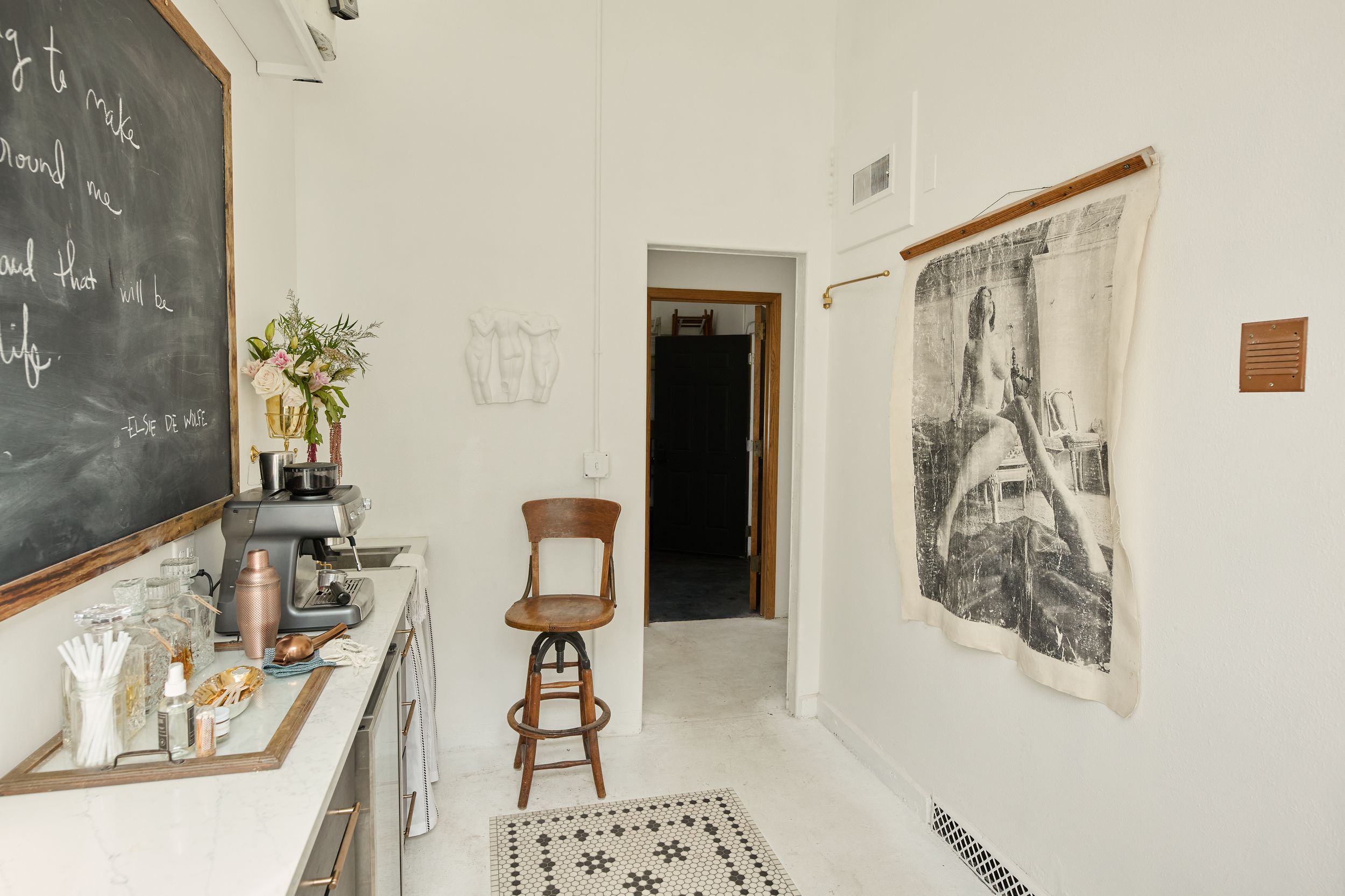 Bright white kitchen nook with chalkboard wall, wooden bar stool, patterned tile rug, and hanging black and white photo print.