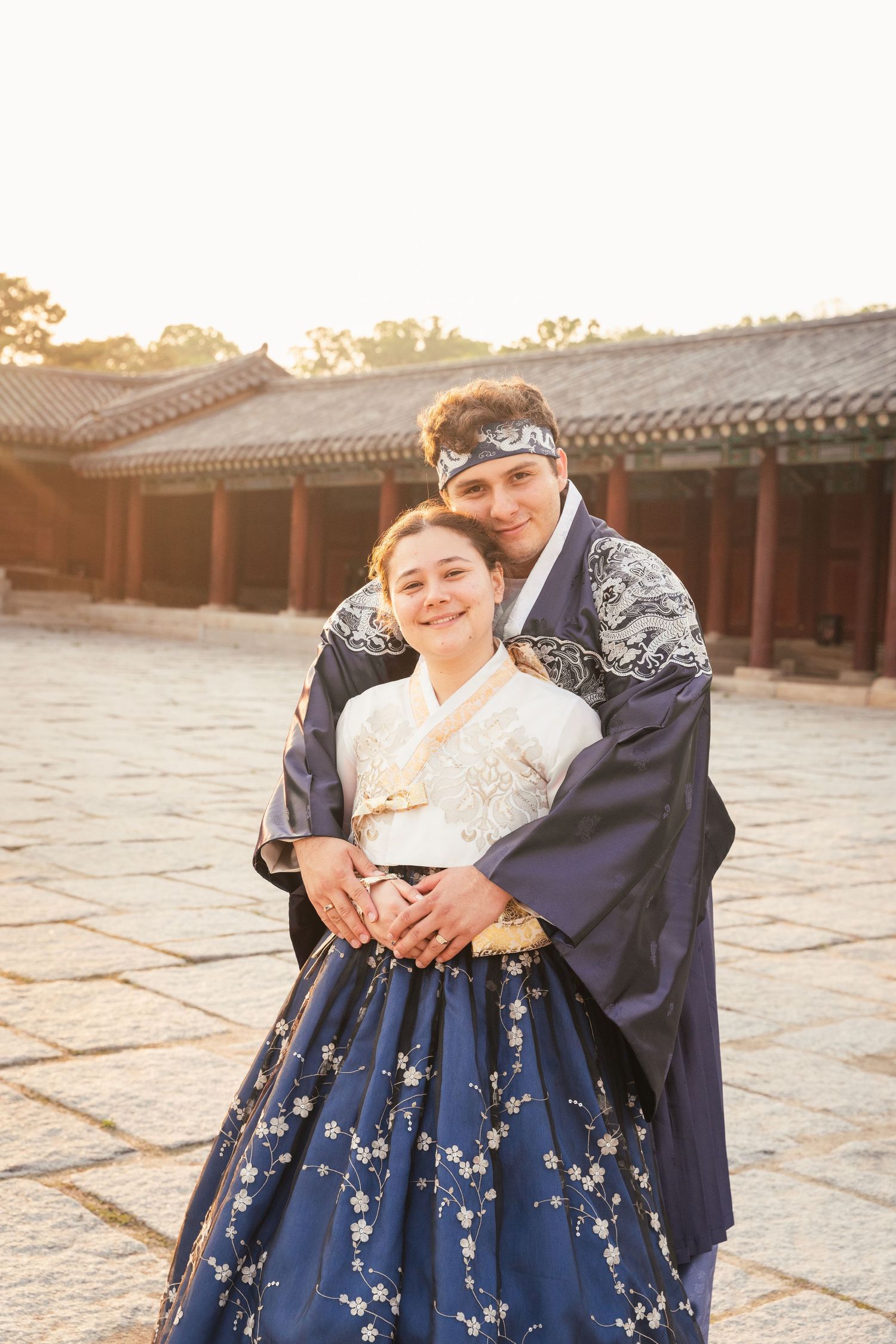 A person wearing a traditional Korean hanbok dress in navy blue with floral embroidery poses in a historic courtyard.