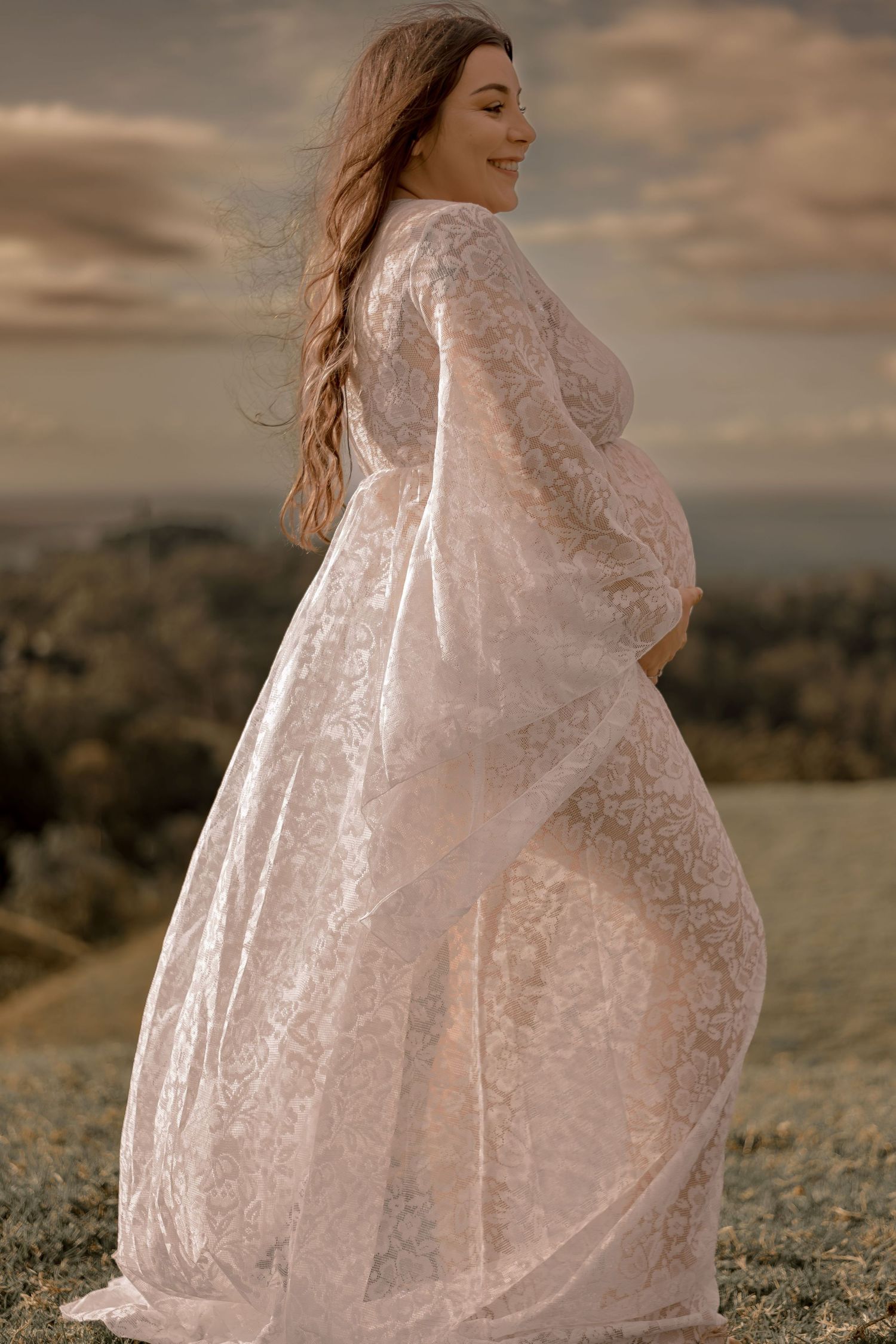 Flowing lace maternity dress catches the wind during golden hour photo session on a hilltop.