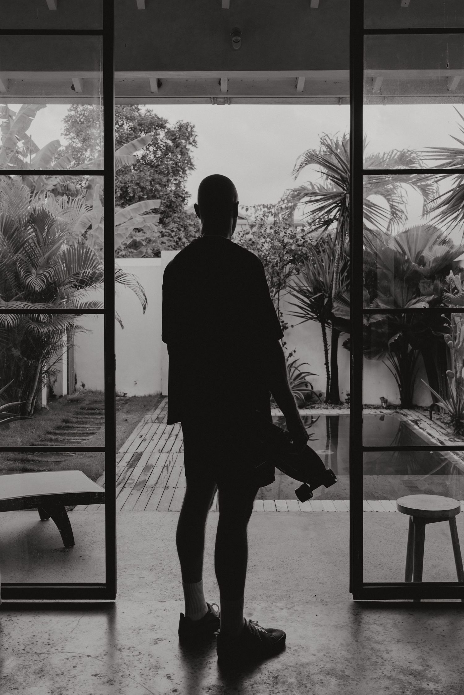 Silhouetted figure stands against glass windows overlooking tropical garden in black and white artistic sequence.