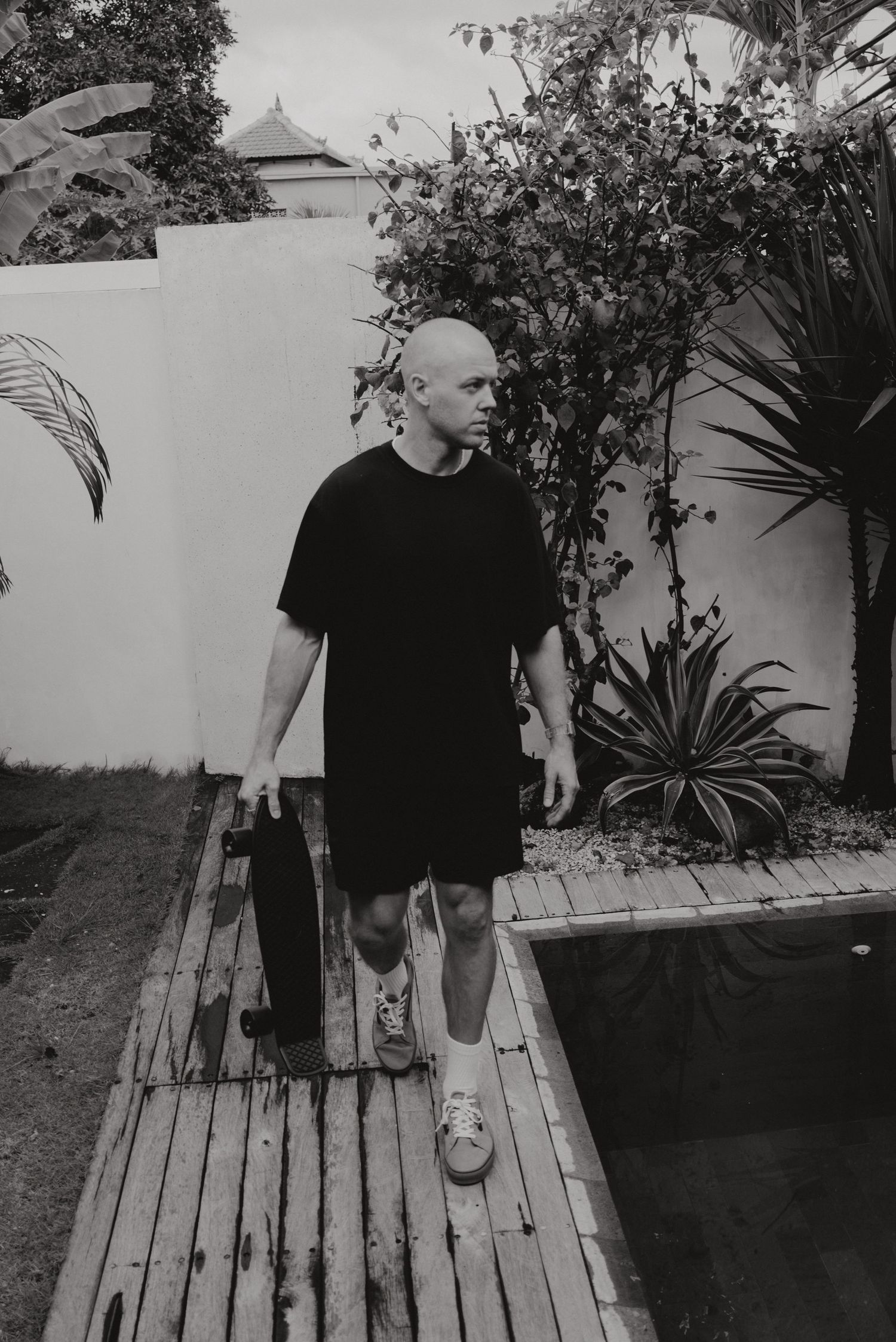 Black and white photo of someone in dark clothing walking on wooden deck near tropical plants and white wall.