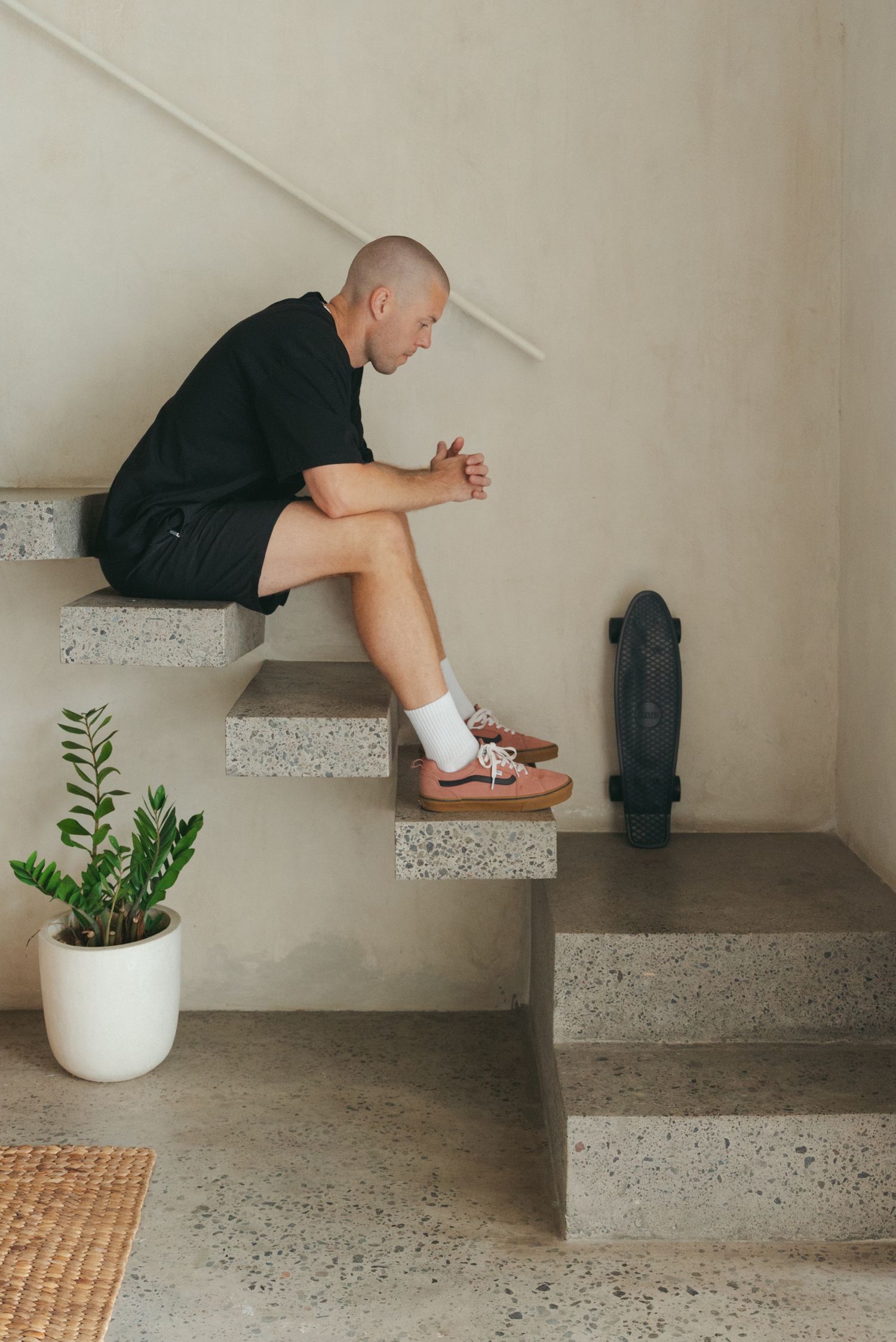 Person sitting on concrete stairs with a skateboard and potted plant in minimalist interior setting.