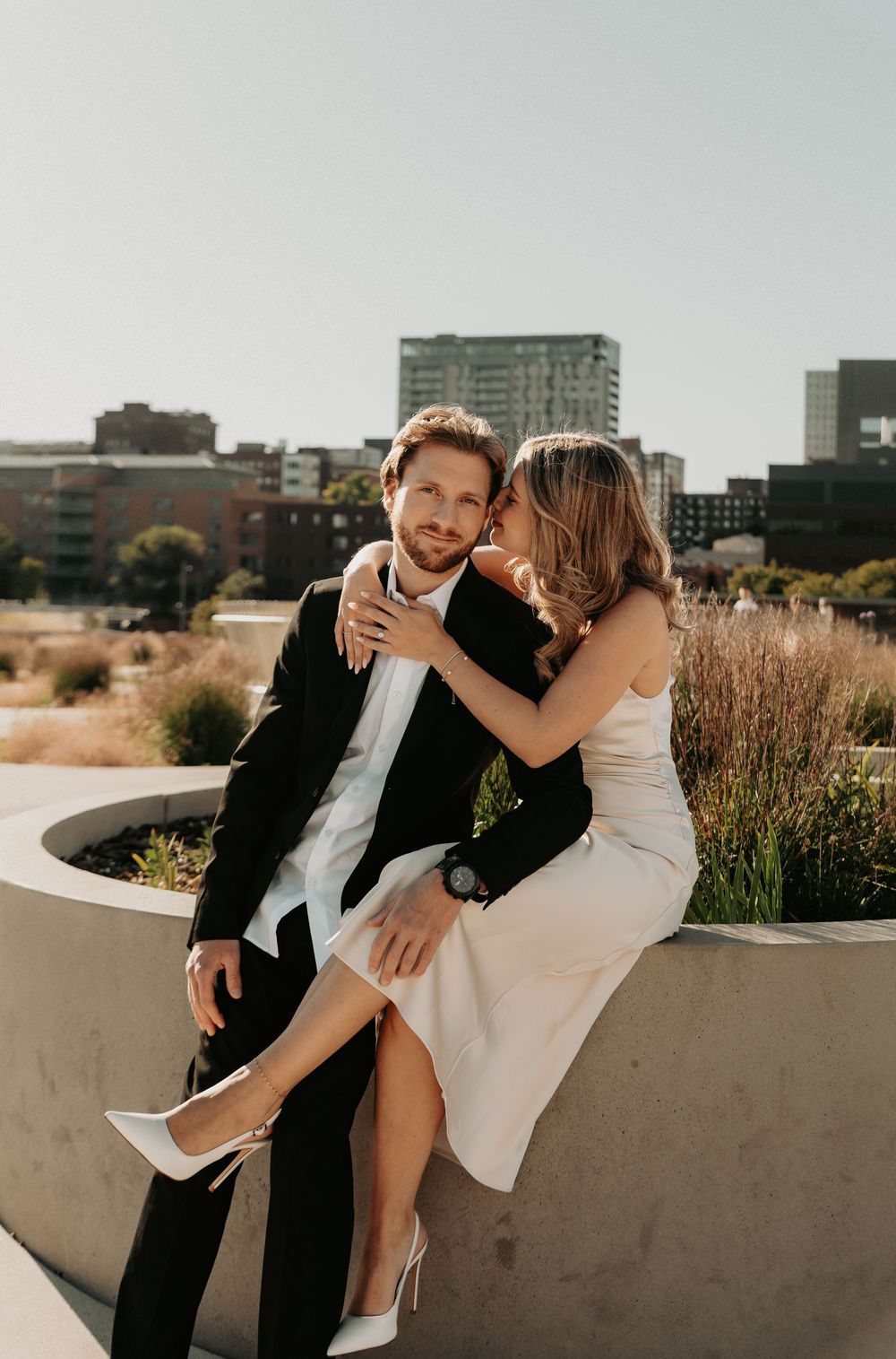 A couple in formal attire shares an intimate moment on a concrete ledge with a city skyline in the background.