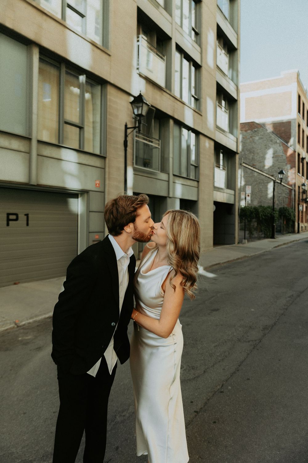 A couple in wedding attire share romantic kisses against an urban backdrop of concrete buildings and sloped streets.