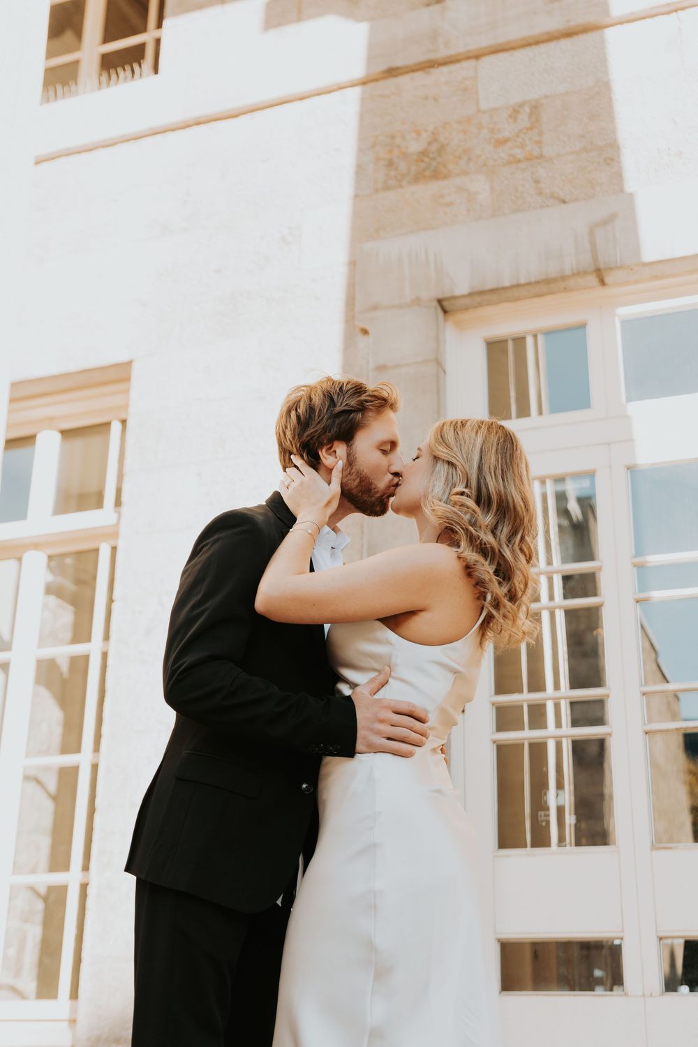 A romantic moment between a couple in wedding attire embracing outside a white building with large windows.