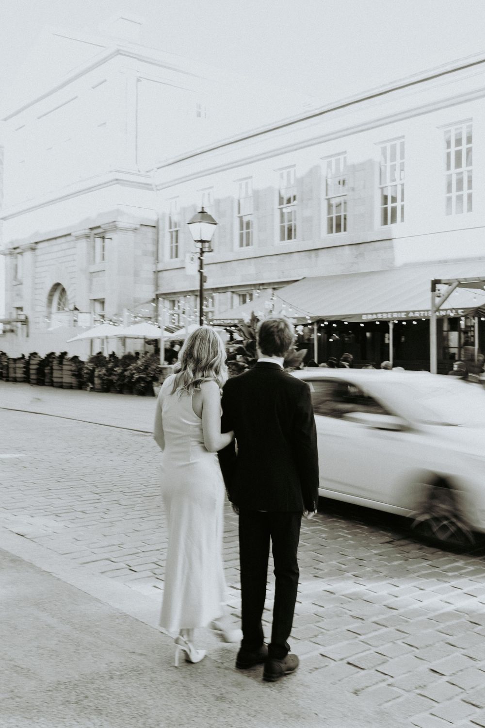 A couple in formal attire walks along a cobblestone street in a vintage black and white photograph series.
