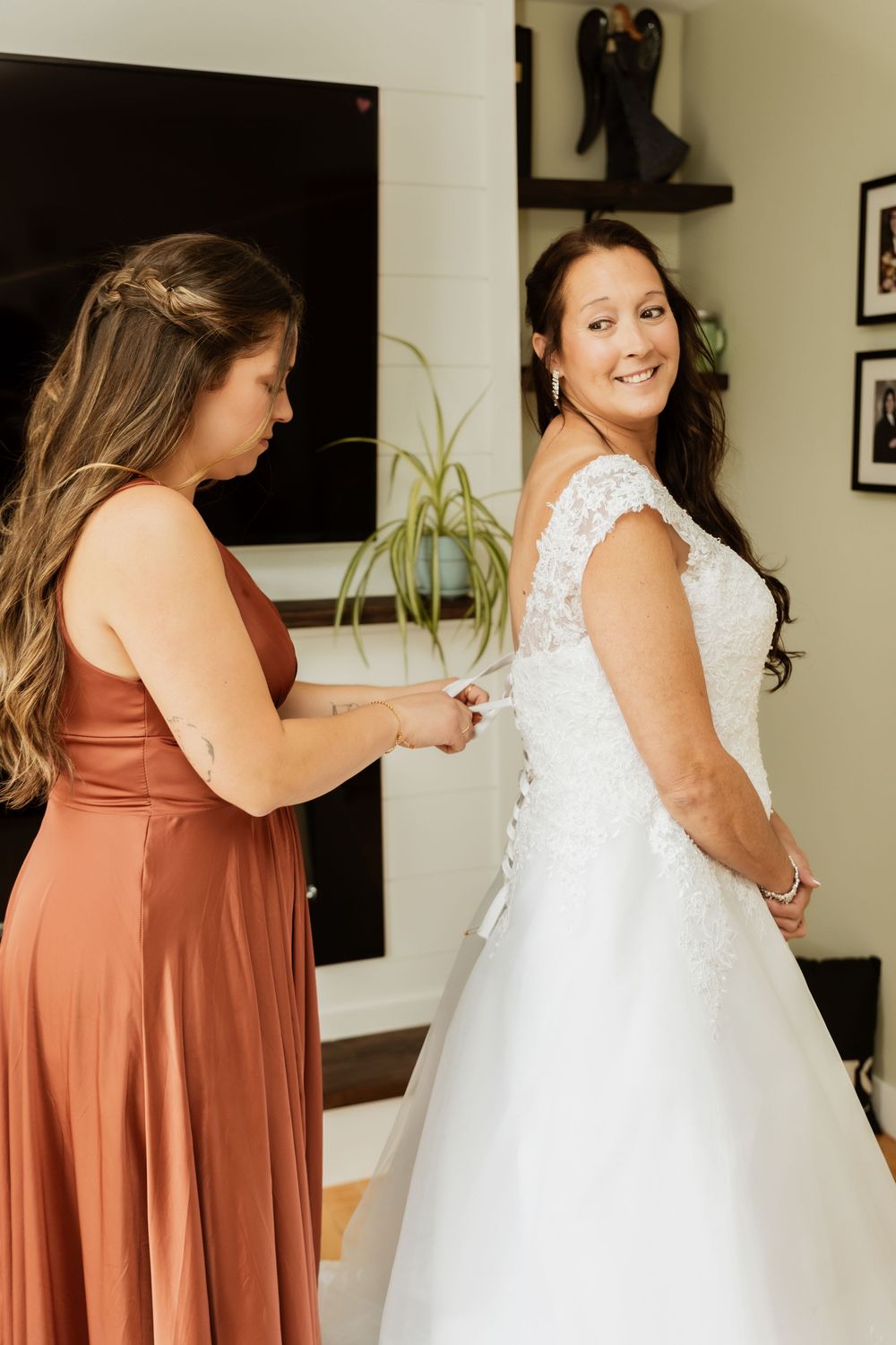 A group of women in terracotta bridesmaid dresses help button up a white wedding gown during pre-ceremony preparations.