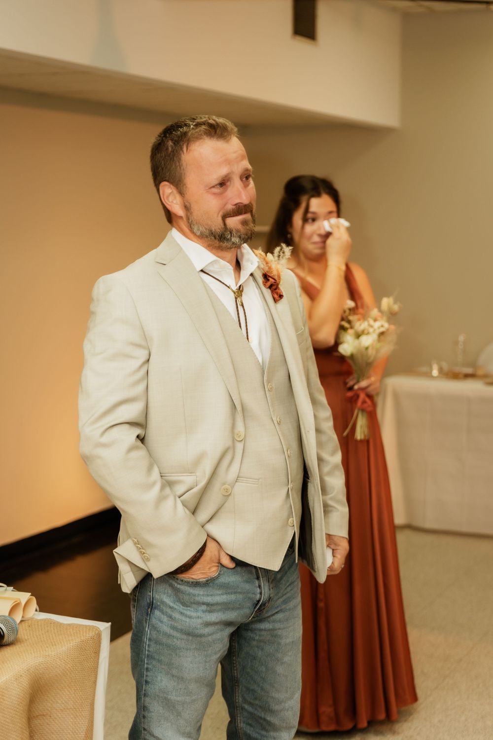 Wedding attendee in light beige suit jacket and jeans stands casually during celebration while bridesmaid looks on.