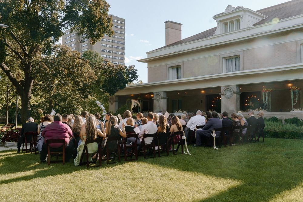 Wedding celebration on the lawn of an elegant white mansion with guests gathered outdoors at sunset.