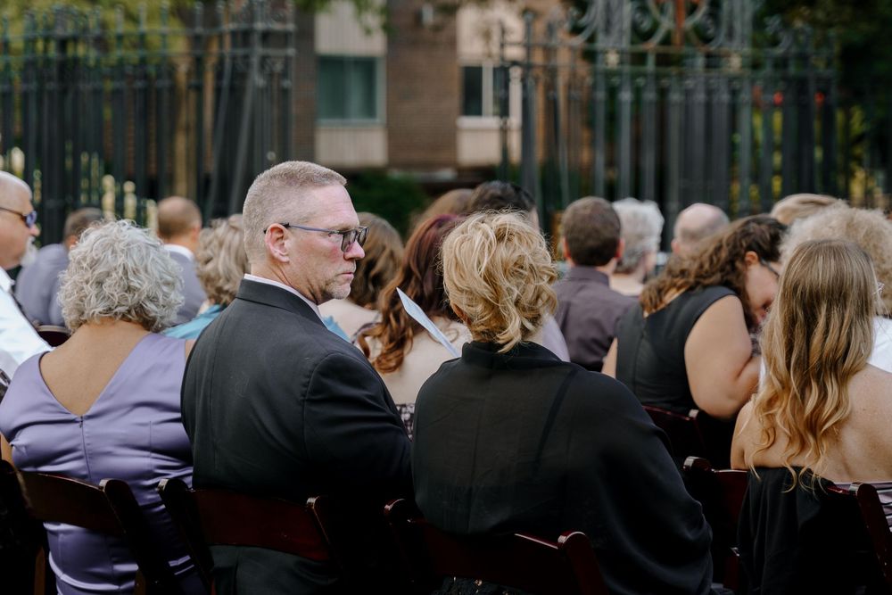 A crowd of people seated outdoors at a formal event with ornate metal gates visible in the background.