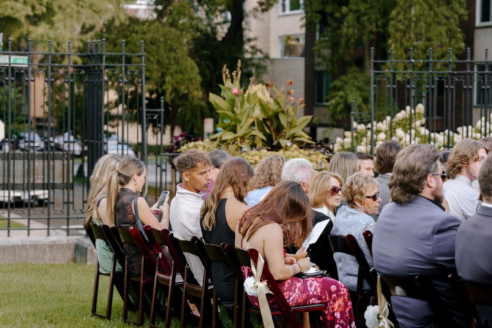A row of outdoor wedding guests seated in white chairs on a sunny day with trees and buildings in the background.