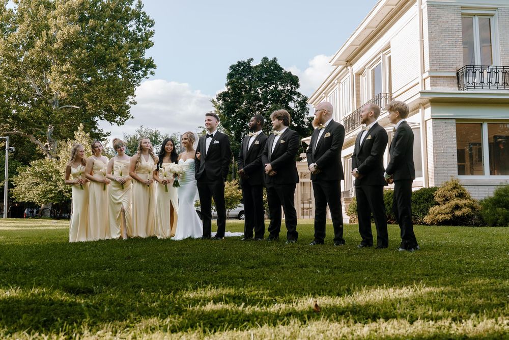Wedding party members stand in a line on a grassy hill outside a white building on a sunny day.