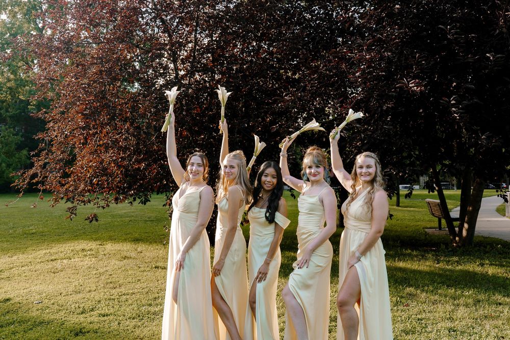 Group in matching blush pink dresses standing together under autumn trees on a grassy hill.