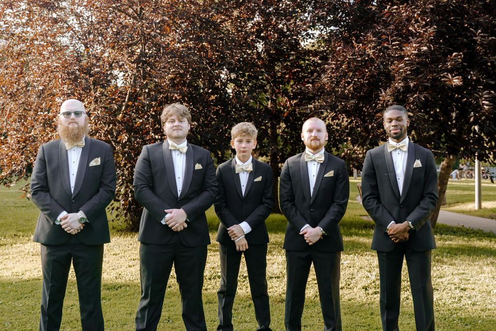 Wedding party members in dark suits stand together outdoors on a sunny autumn day with fall foliage in background.