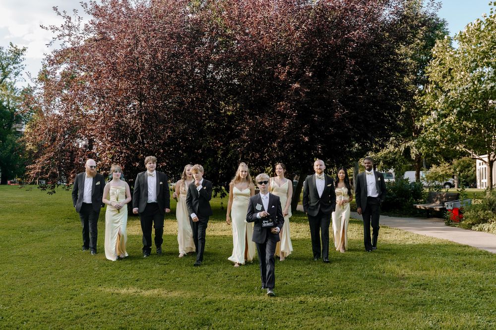 Wedding party stands in a row under pink cherry blossom trees in a lush garden setting during spring.