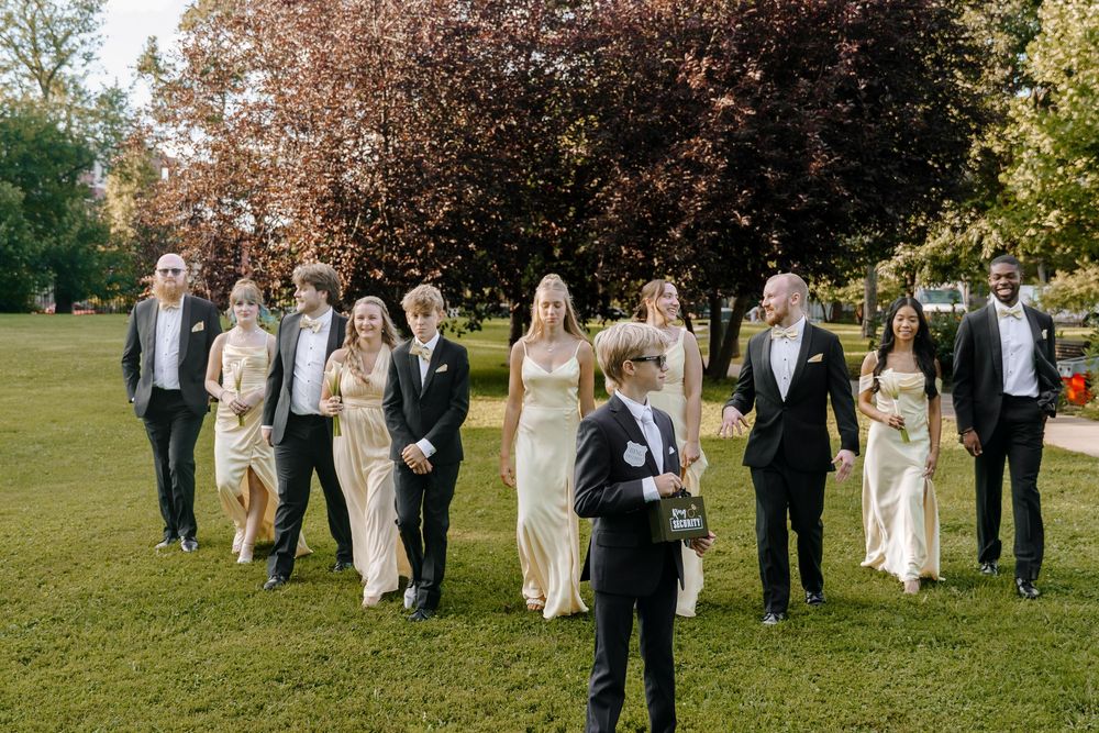 Wedding party members walking in a line across a sunny green lawn during formal photographs.