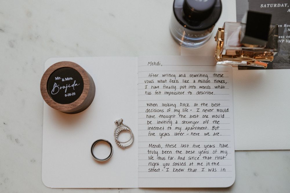 Wedding detail flatlay with perfume bottles, rings, and invitation cards on white marble surface.