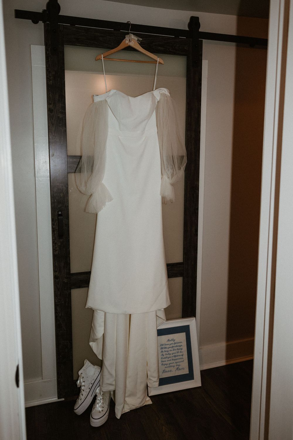 White wedding attire including a dress and jumpsuit hanging on wooden hangers against a dark wall.