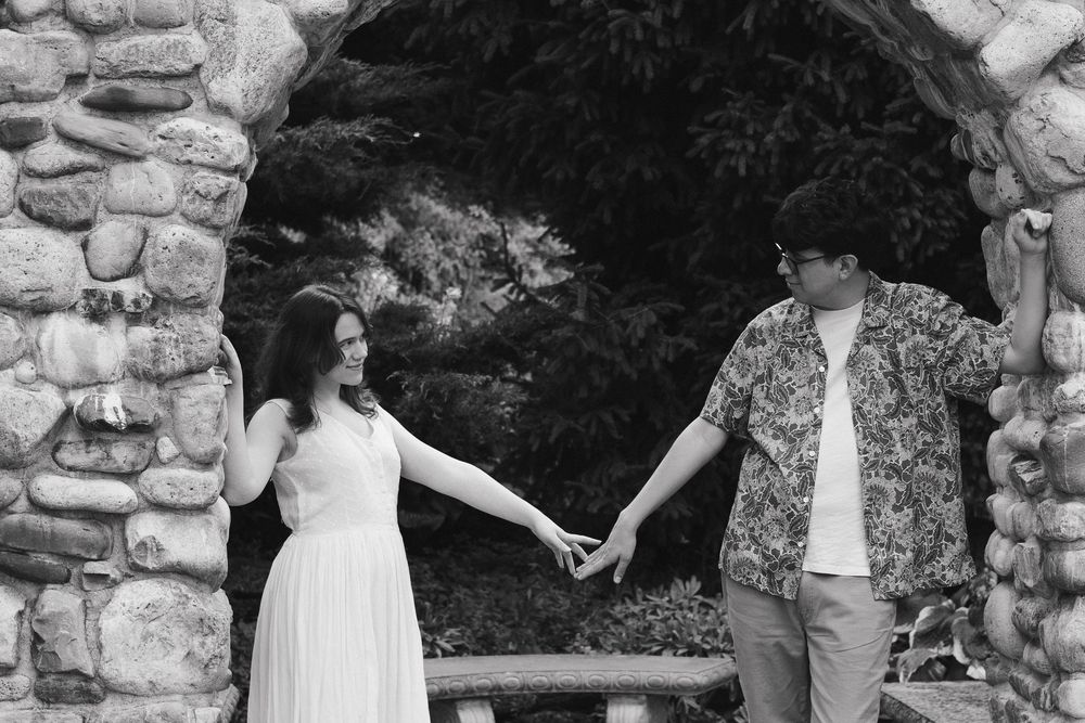 A couple holds hands while standing beneath a rustic stone archway in black and white photography.