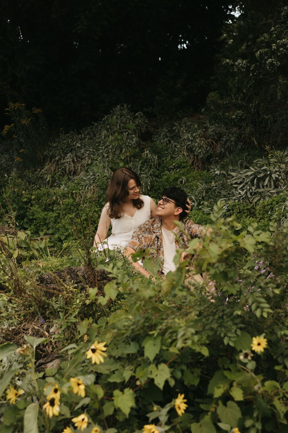 Couple sitting together among lush greenery and yellow wildflowers in a romantic outdoor garden setting.