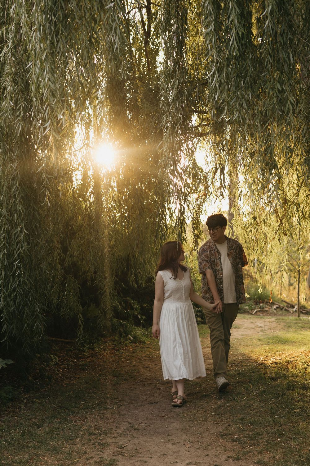 A couple walks hand in hand beneath golden sunlit willow trees along a serene garden path.