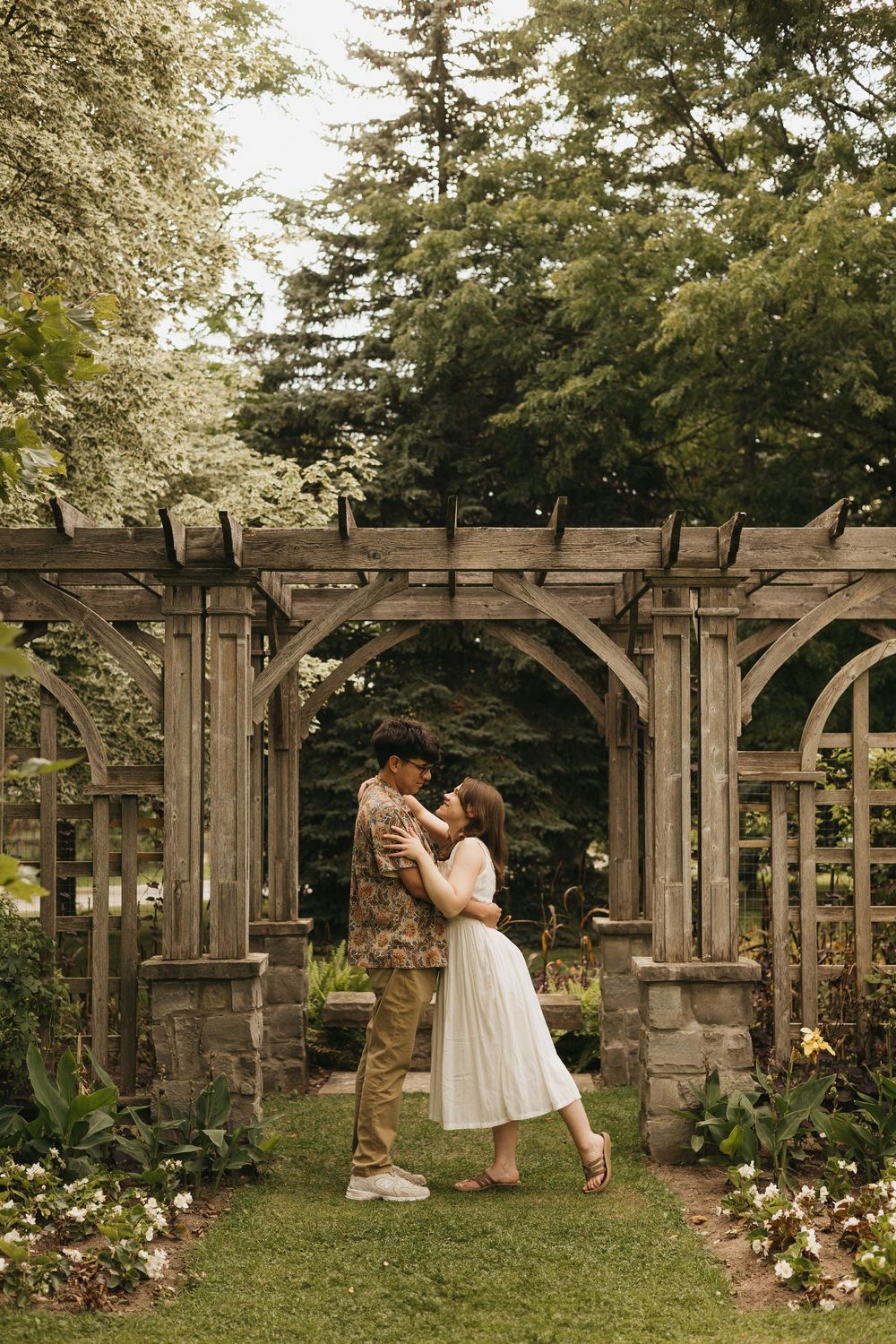 A couple shares romantic moments under a wooden garden pergola surrounded by lush greenery and blooming flowers.