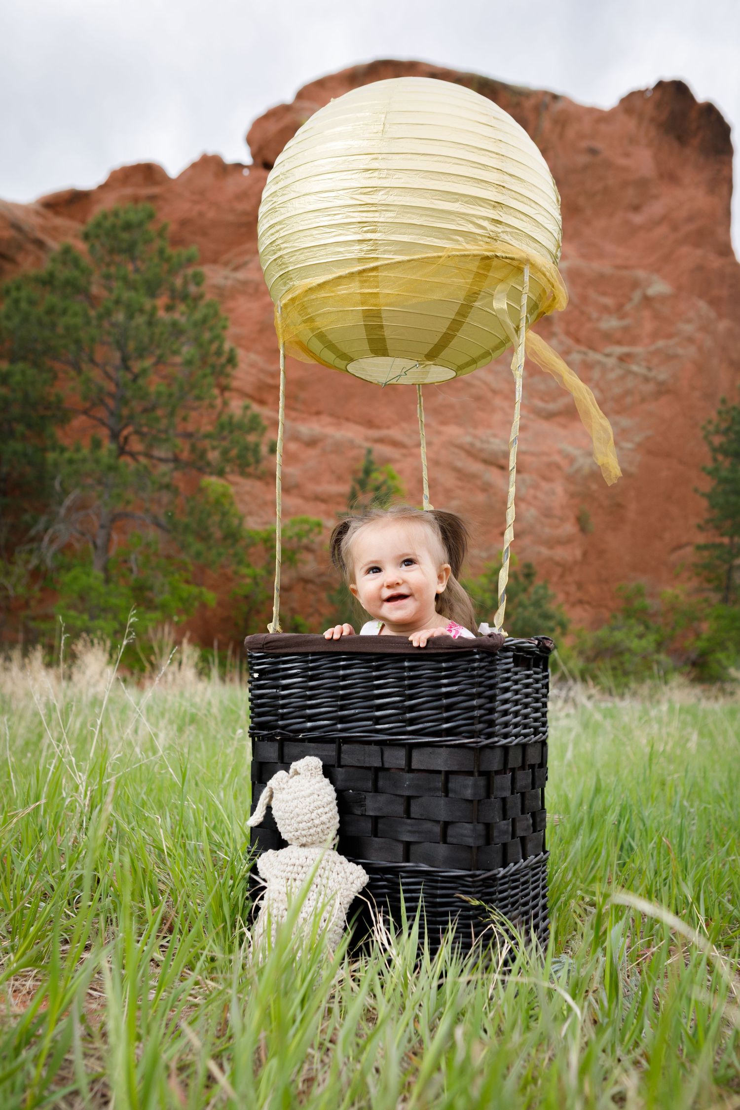 A baby sitting in a black wicker basket against a red rock formation and grassy field.