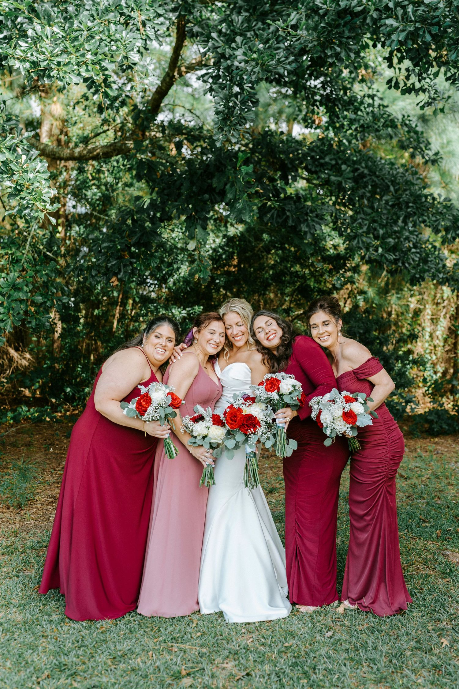 Wedding party in burgundy bridesmaid dresses and white bridal gown pose playfully in a garden setting.