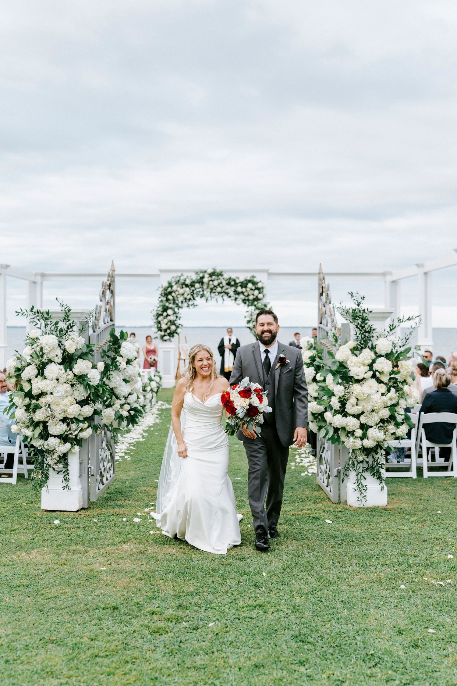 Outdoor wedding ceremony setup with white floral arrangements lining the aisle on a grassy hill overlooking the water.