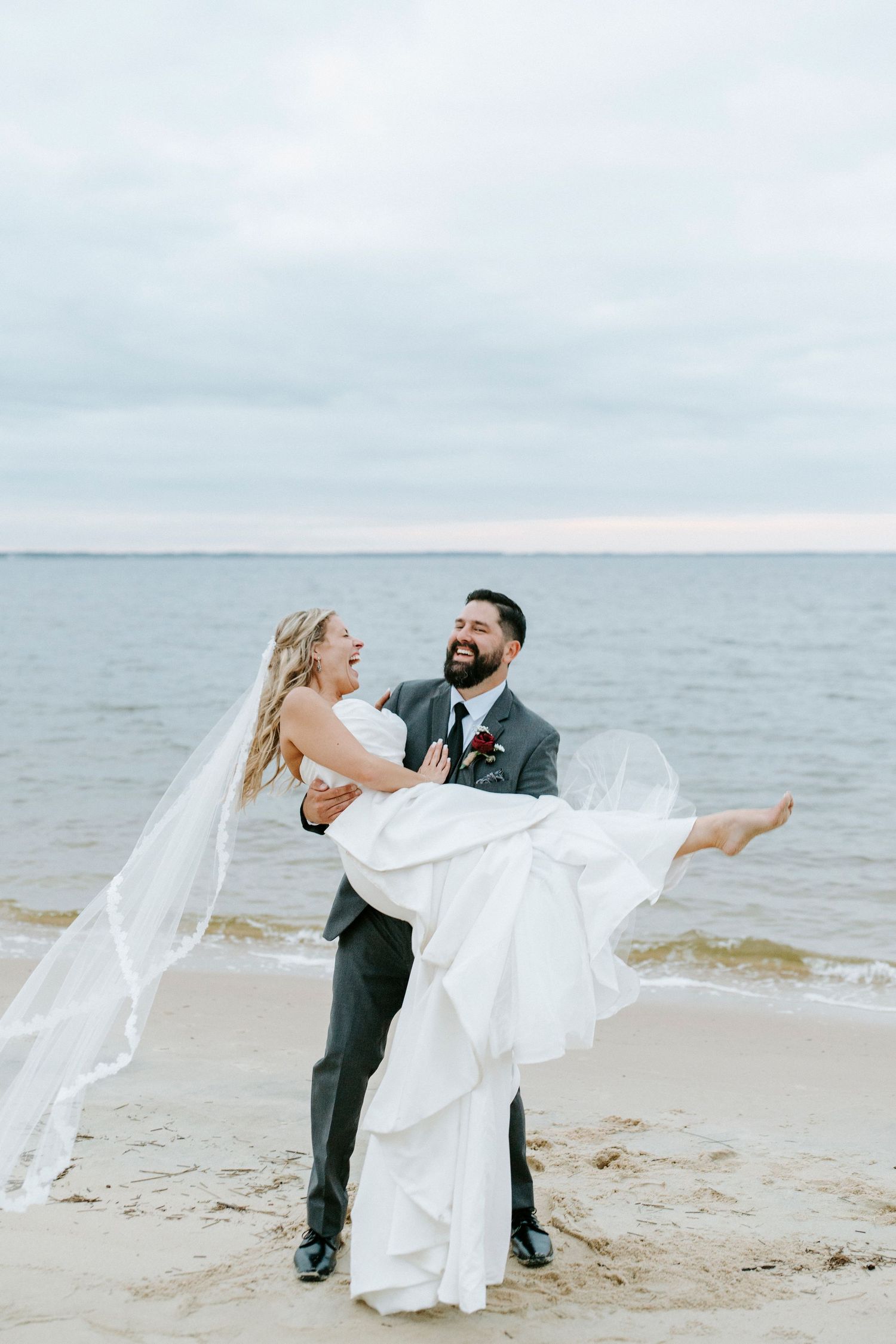 Newlyweds dance playfully on a beach as the bride's white dress flows in the ocean breeze.