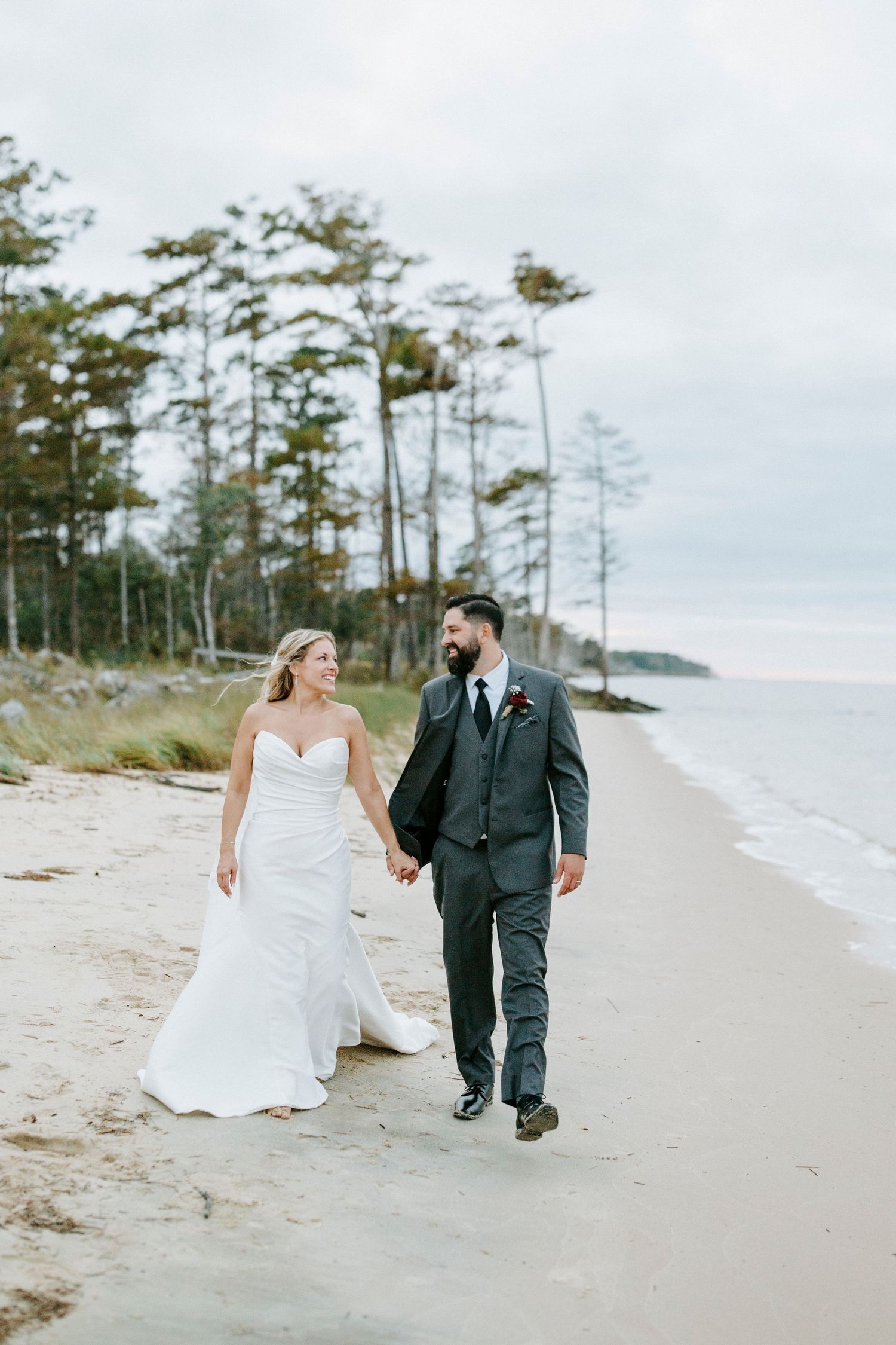 Playful couple in wedding attire running along a scenic coastal beach path.