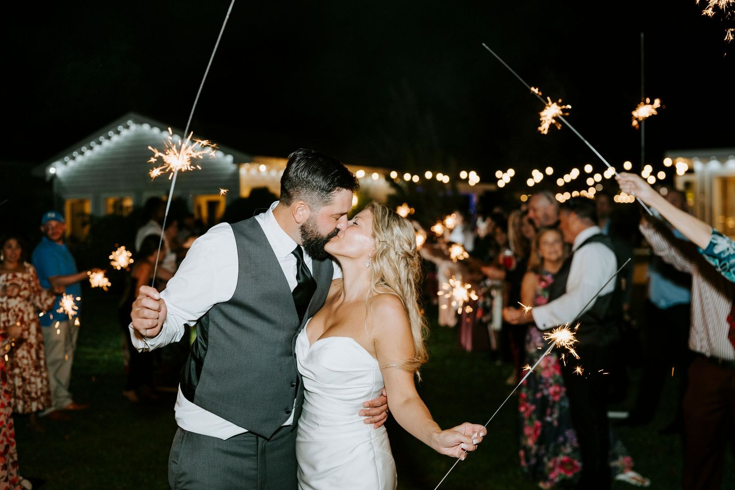 Wedding guests hold sparklers creating a magical light tunnel at an outdoor nighttime celebration.