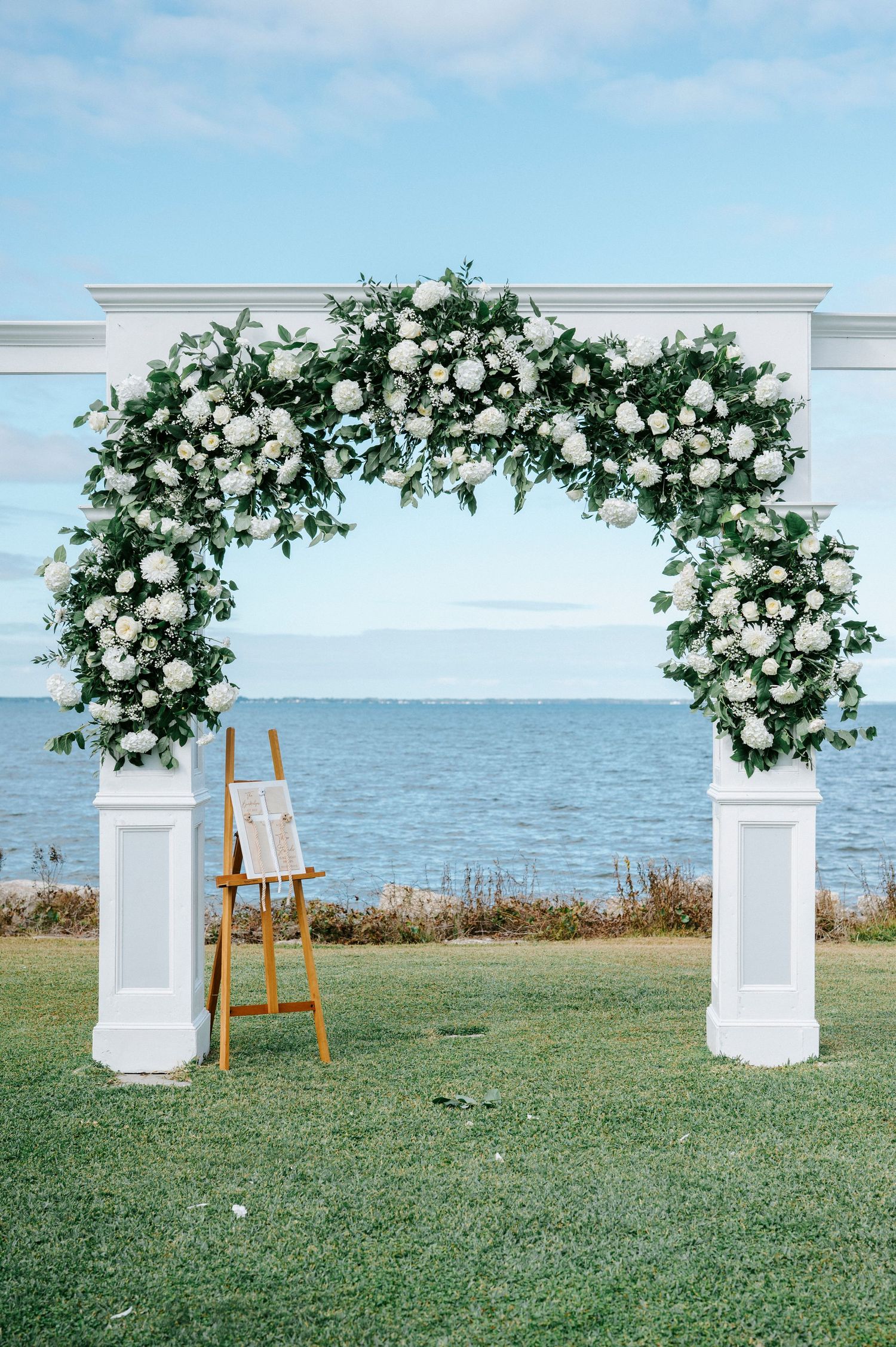 A stunning white floral wedding arch overlooking the ocean with an easel stand on a grassy lawn.