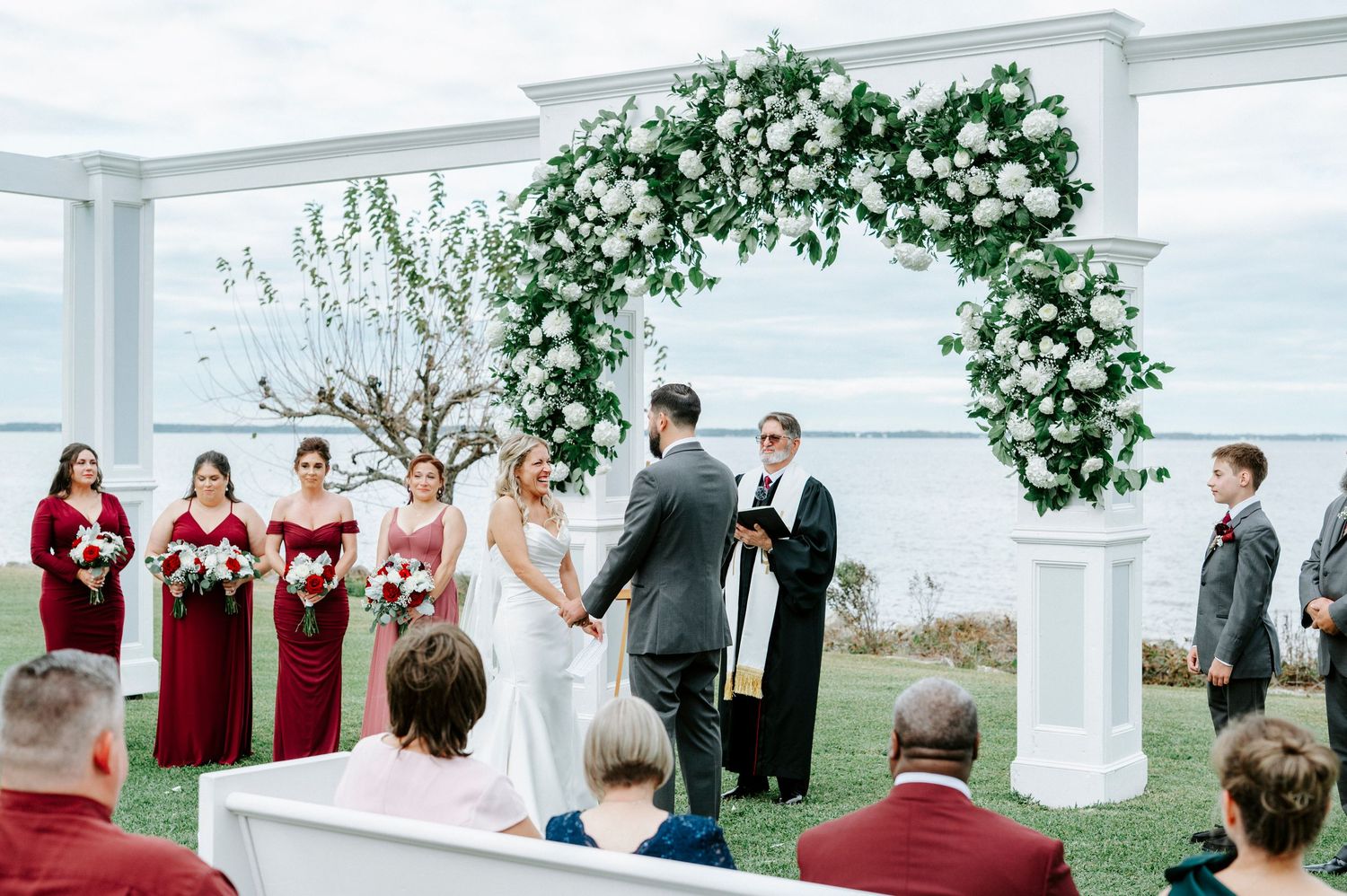 Outdoor wedding ceremony with white pergola and red-dressed attendants in a garden setting.