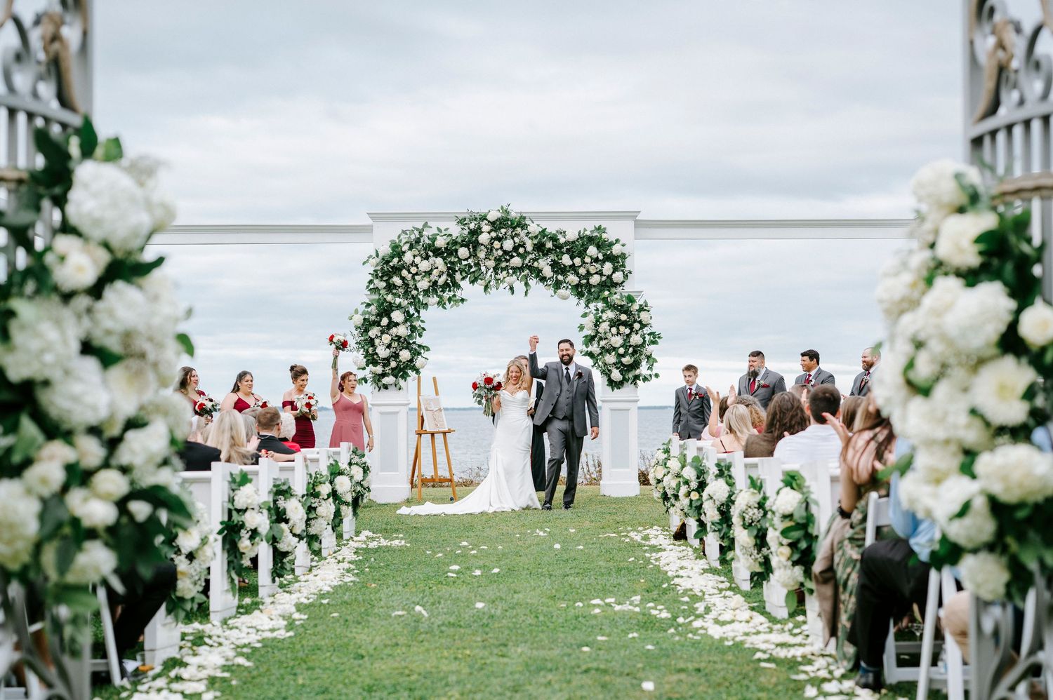 Outdoor wedding ceremony setup with white floral arch and guests seated on white chairs by the water.