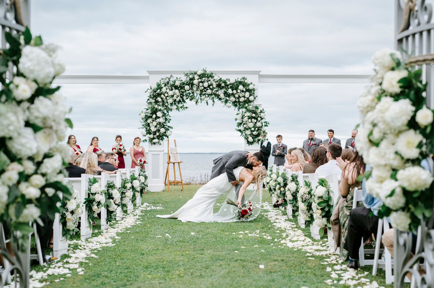 Wedding ceremony setup with white floral arches and chairs on a manicured lawn overlooking the water.