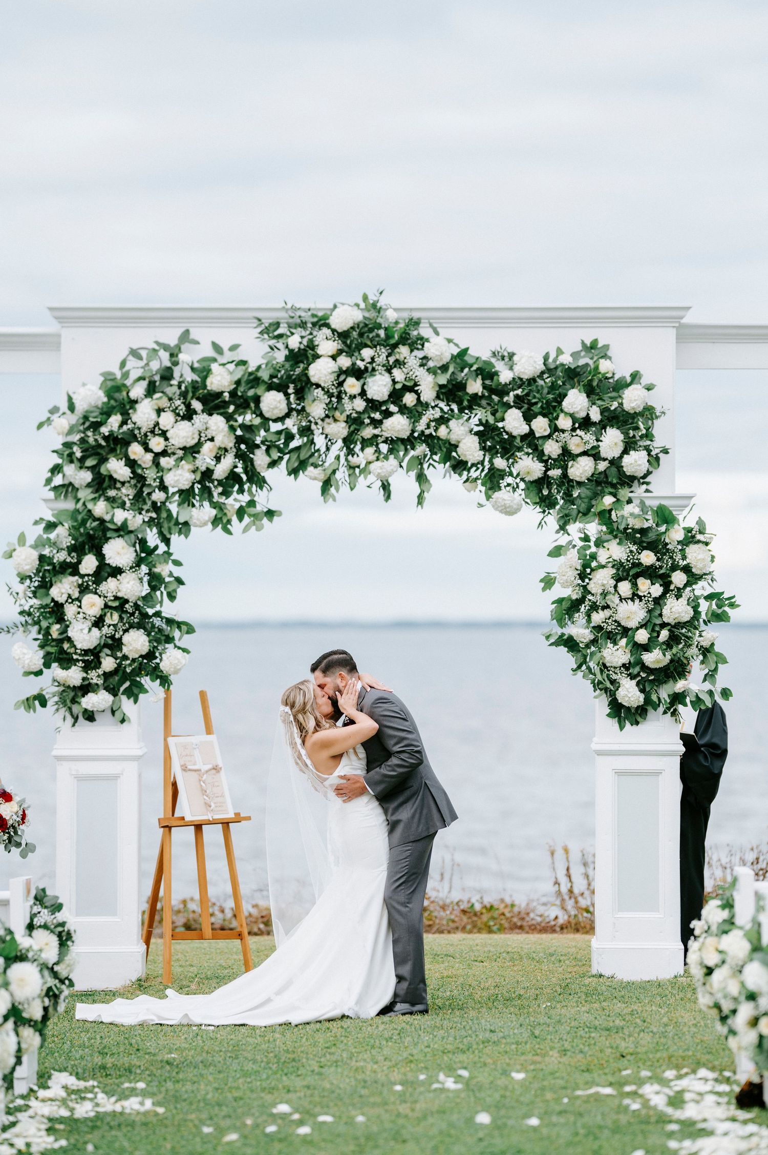 Elegant wedding arch adorned with white roses and greenery creates a romantic backdrop for an oceanside ceremony.